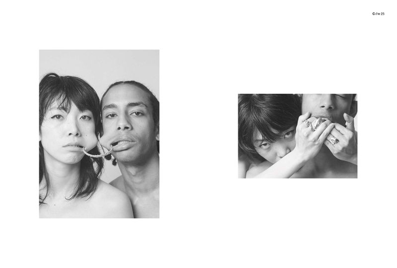 Black and white studio portrait of two people with dark hair. One has jewelry between their lips and the other covers another's mouth.
