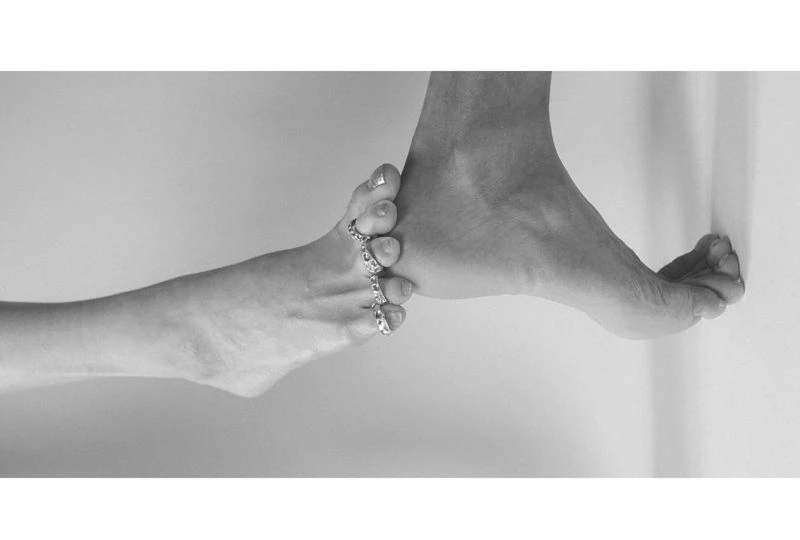 Monochrome image showing two intertwined adult feet, one adorned with toe rings, set against a neutral wall background in a studio setting.