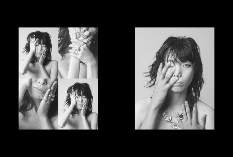 Black and white fashion shot of a woman with short dark hair wearing rings, covering one eye with her hand, and a necklace.