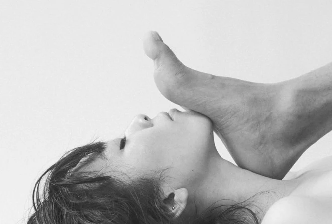 Monochrome portrait of a young woman with dark hair, lying down with a human foot resting on her face, set against a white background.