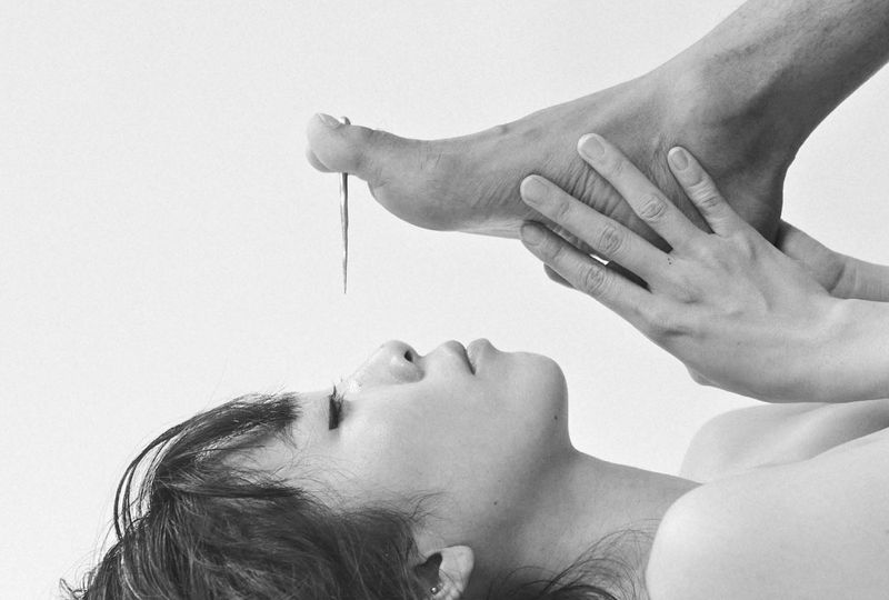 Monochrome photo of a woman lying down with eyes closed, a foot with a nail piercing the toe above her face, held by a hand.