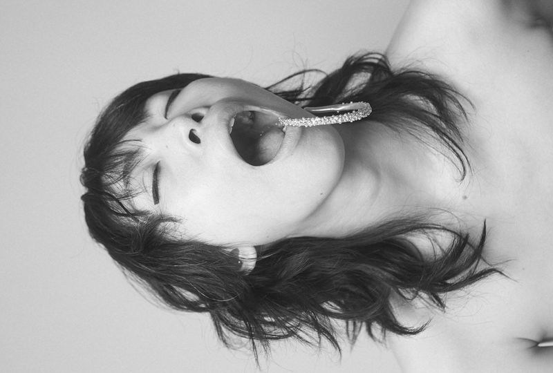 Artistic black and white portrait of a woman with dark hair lying down, wearing a sparkly jeweled bracelet in her mouth.
