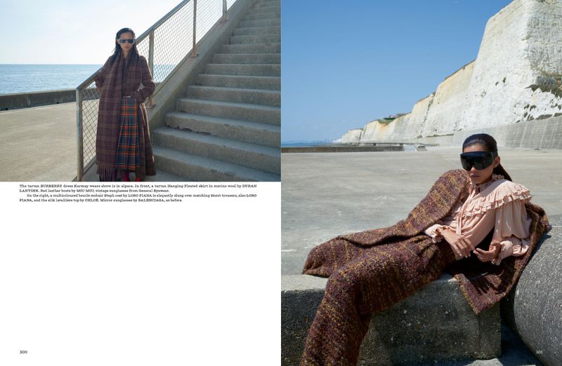 Fashion editorial shot featuring a woman in sunglasses, a long coat, and a plaid skirt, seated against a white cliff backdrop near the ocean.