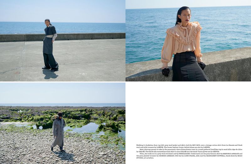 Fashion editorial shot of a model wearing a grey wool jacket, vintage shirt, and black trousers against a seaside backdrop.