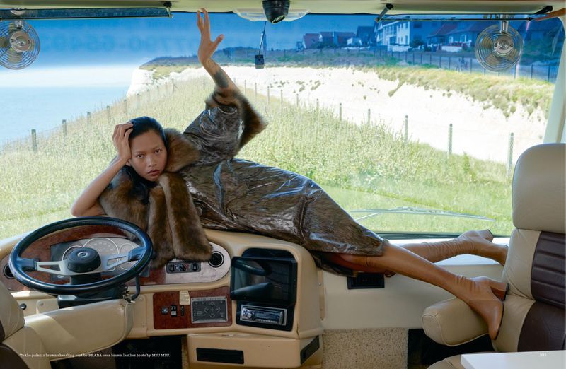 Woman in silver dress and brown fur stole poses inside RV with coastal view. Car dashboard, steering wheel, and seaside vista visible.