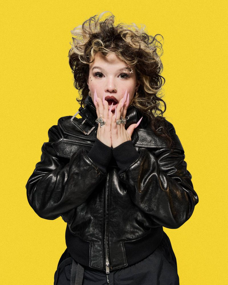 Portrait of a young adult female with curly hair and long nails, wearing a black jacket with a bright yellow background.