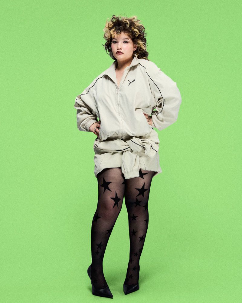 A woman with curly hair poses in a studio wearing a beige jacket and skirt, sheer black patterned tights, and black high heels.