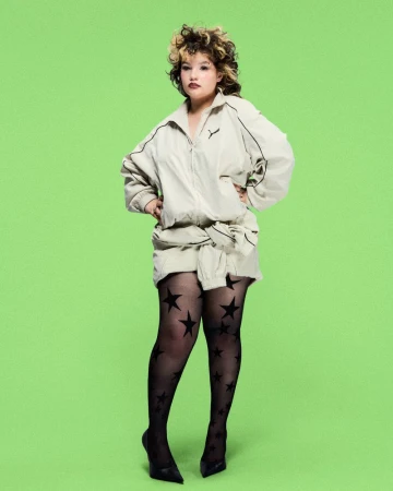 A woman with curly hair poses in a studio wearing a beige jacket and skirt, sheer black patterned tights, and black high heels.