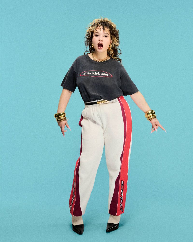 Full-length shot of a stylish adult woman with curly hair in a t-shirt and athletic pants, posing against a bright blue background.