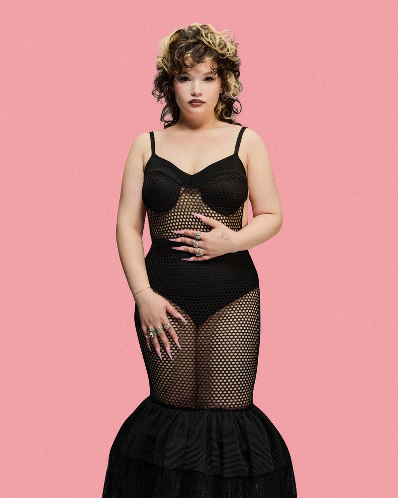 Woman in a black knit dress poses against a pink backdrop, showcasing a blend of fashion and visual artistry in a studio setting.