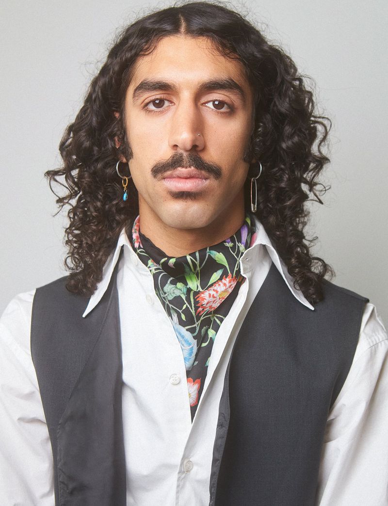 Portrait of a dark-haired adult male with a mustache wearing a floral ascot, black vest, and white shirt against a white background.