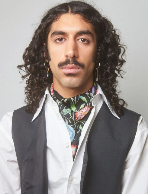 Portrait of a dark-haired adult male with a mustache wearing a floral ascot, black vest, and white shirt against a white background.