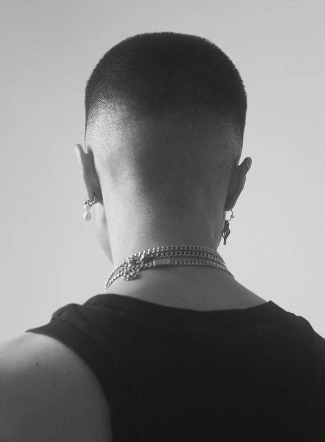 Back view of a person with a buzzcut wearing a black tank top and silver necklaces, photographed in a monochromatic studio setting.