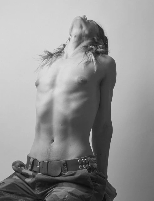 Monochromatic portrait of a shirtless person in camo pants, looking up with pigtails, captured in a studio setting with even lighting.