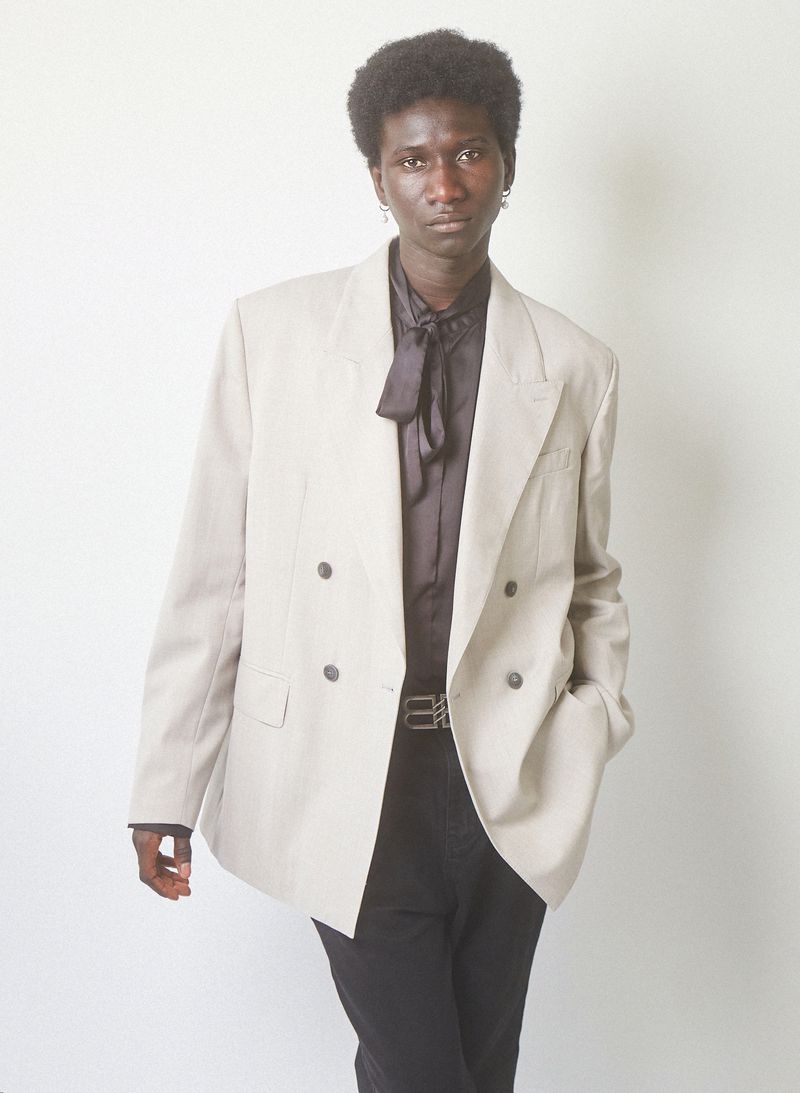 Young adult male in beige suit and black shirt poses in front of a white backdrop with neutral expression. Fashion editorial shot.
