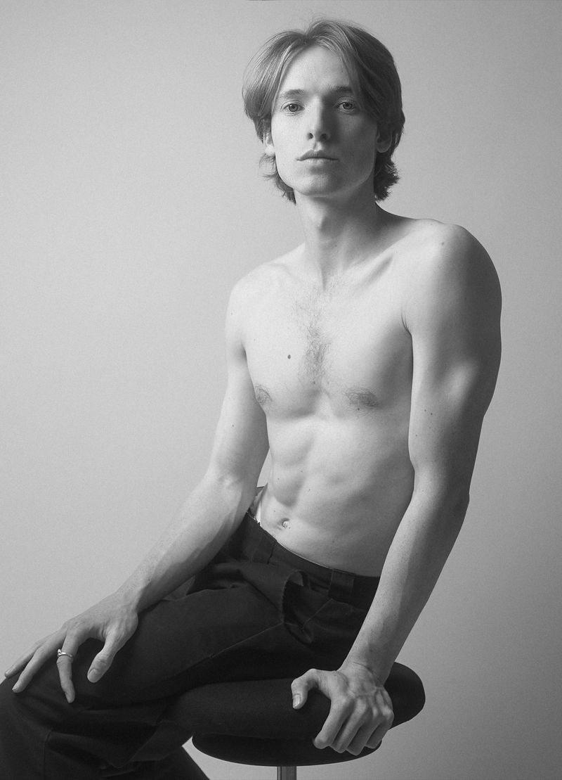Model Karl Heinz Hirschberger sitting shirtless on a stool wearing black trousers in a black and white studio portrait.