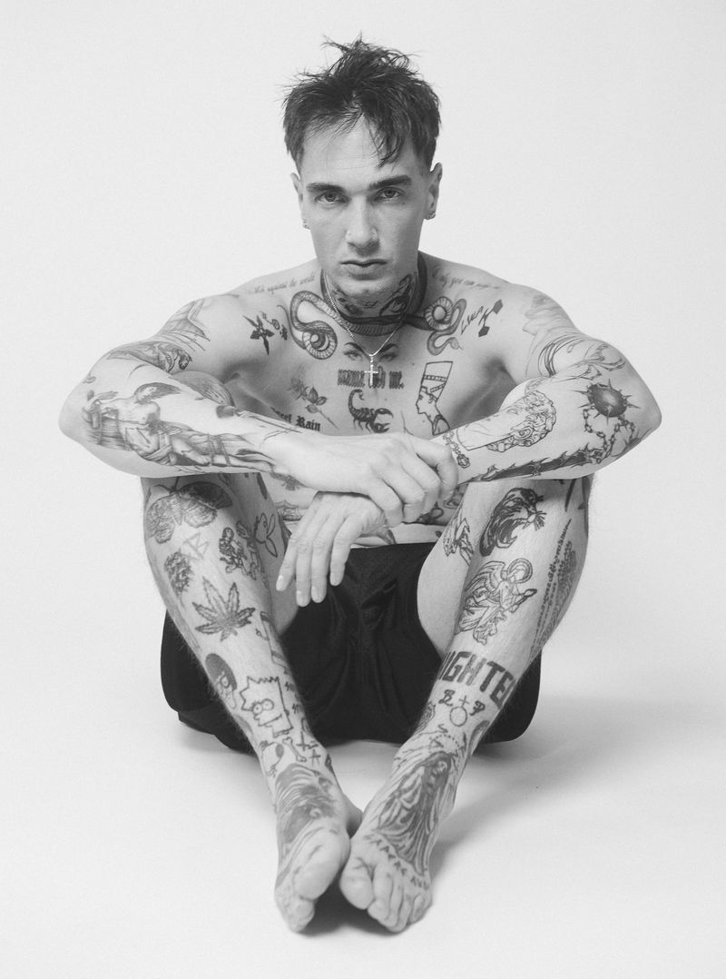 Black and white portrait of a tattooed adult male seated with his arms resting on his knees in a photo studio on a seamless backdrop.
