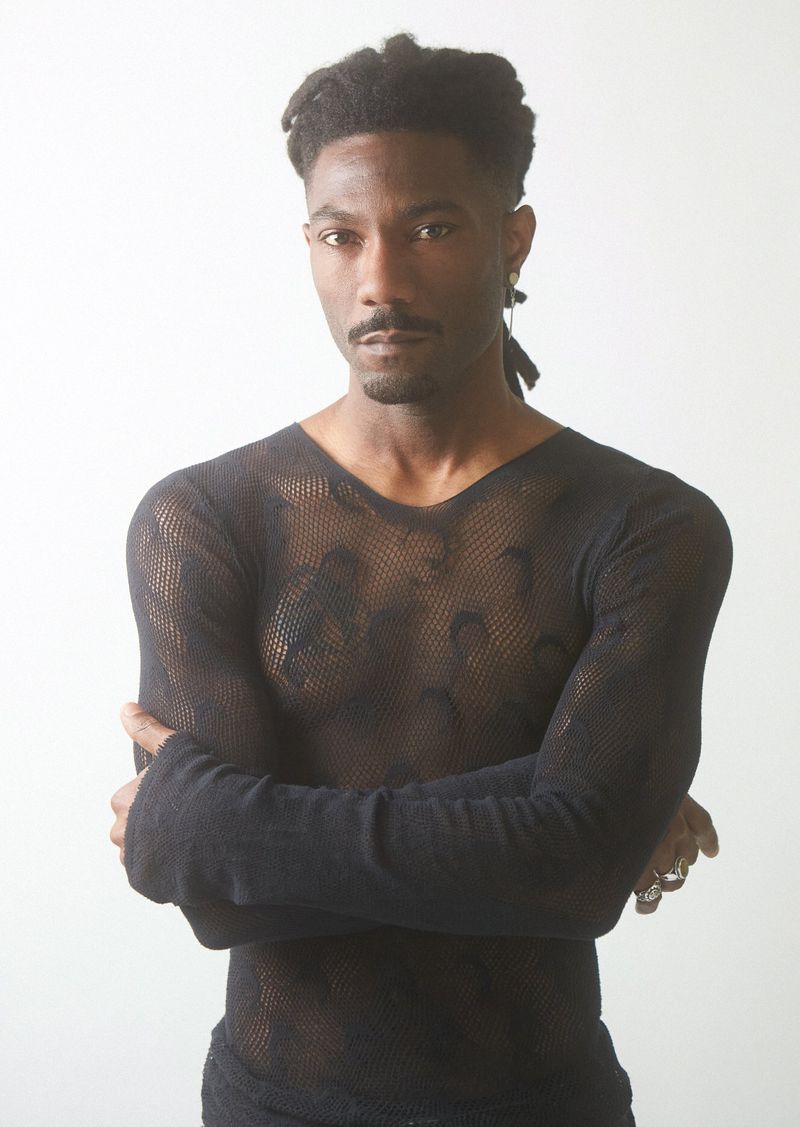 Portrait of adult male wearing a black, long-sleeved mesh shirt, standing indoors against a white background with arms crossed.