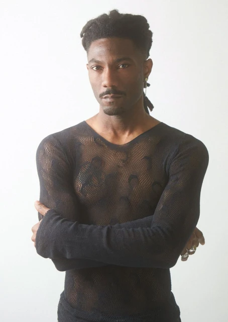 Portrait of adult male wearing a black, long-sleeved mesh shirt, standing indoors against a white background with arms crossed.