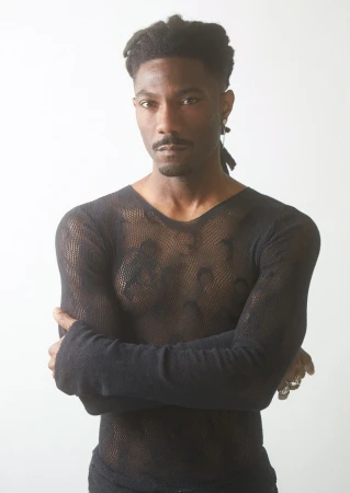 Portrait of adult male wearing a black, long-sleeved mesh shirt, standing indoors against a white background with arms crossed.