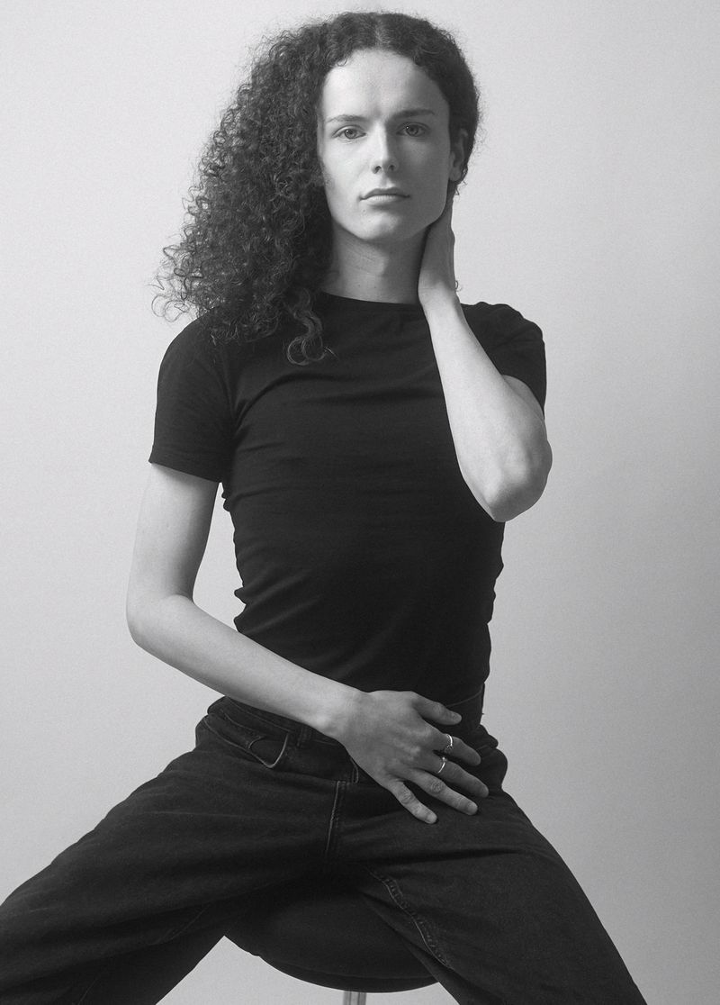 Black-and-white portrait of a young adult with curly hair, wearing a black t-shirt and dark pants, seated against a plain background.