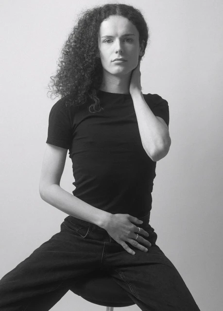 Black-and-white portrait of a young adult with curly hair, wearing a black t-shirt and dark pants, seated against a plain background.