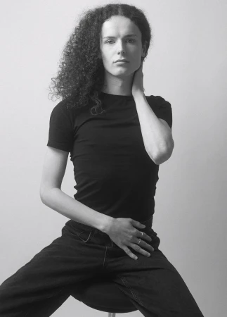 Black-and-white portrait of a young adult with curly hair, wearing a black t-shirt and dark pants, seated against a plain background.