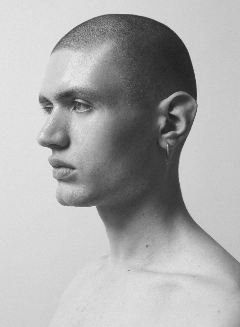 Striking black and white portrait of a young adult male with short hair, showcasing his profile with a minimal earring against a plain background.