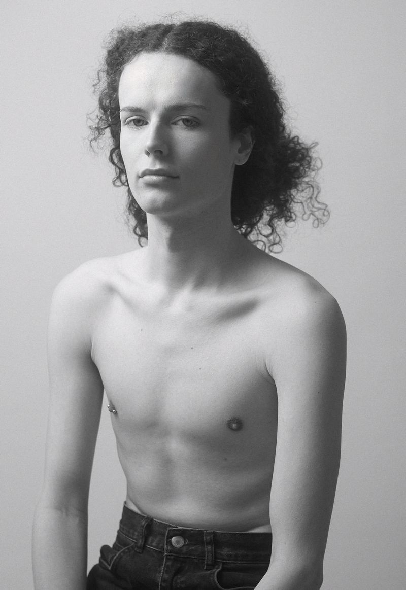 Monochrome portrait of a young adult with curly hair, bare torso, and dark denim jeans against a light background.