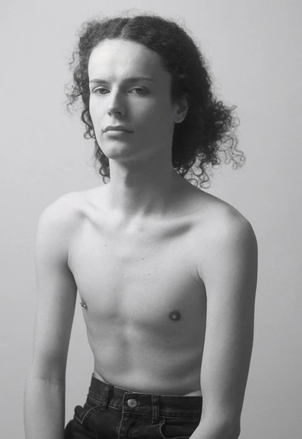 Monochrome portrait of a young adult with curly hair, bare torso, and dark denim jeans against a light background.