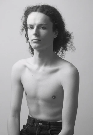Monochrome portrait of a young adult with curly hair, bare torso, and dark denim jeans against a light background.