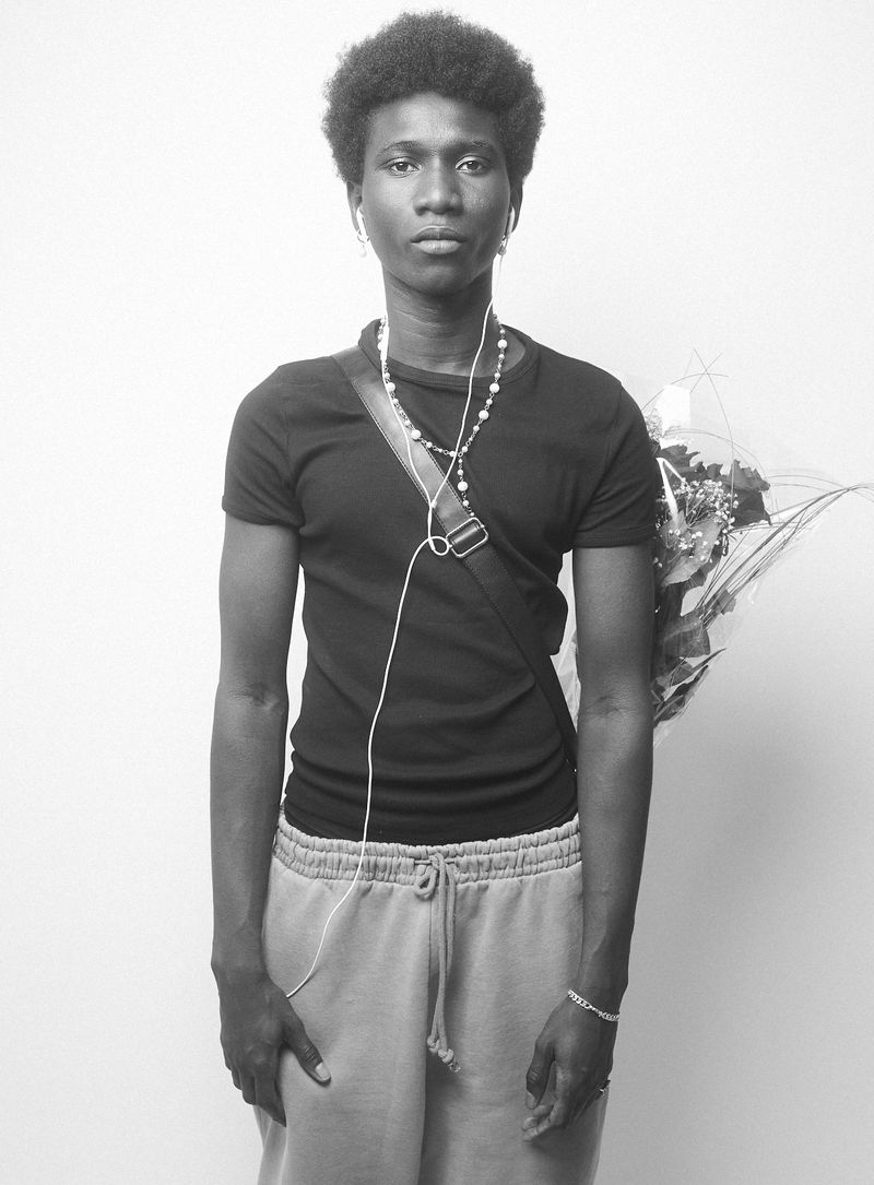 Monochromatic portrait of a young adult with curly hair, wearing a T-shirt, sweatpants, beads, and carrying flowers in a studio setting.