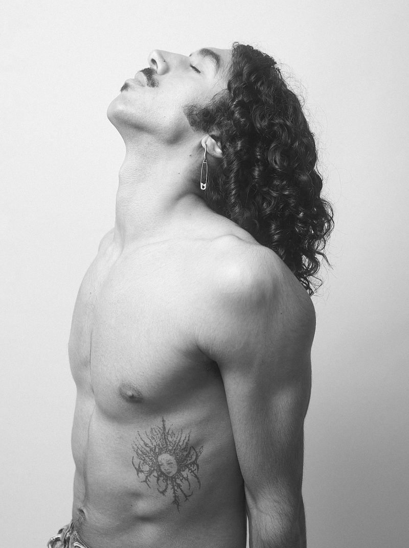 Black and white side profile portrait of a shirtless man with curly hair and a safety pin earring against a plain backdrop.