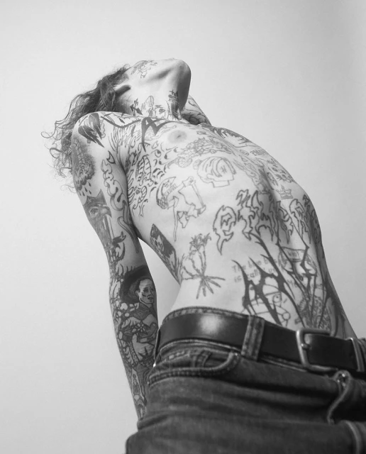 Artistic black and white studio shot of a shirtless adult with tattoos, wearing jeans and a belt, bending backward with head tilted back.