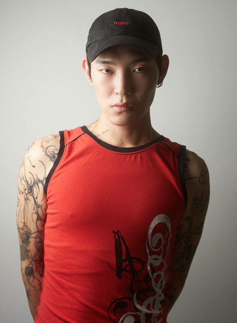 Portrait of a young adult male in a red tank top and black cap, facing the camera, showcasing tattoos in a neutral indoor environment.