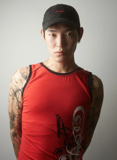 Portrait of a young adult male in a red tank top and black cap, facing the camera, showcasing tattoos in a neutral indoor environment.