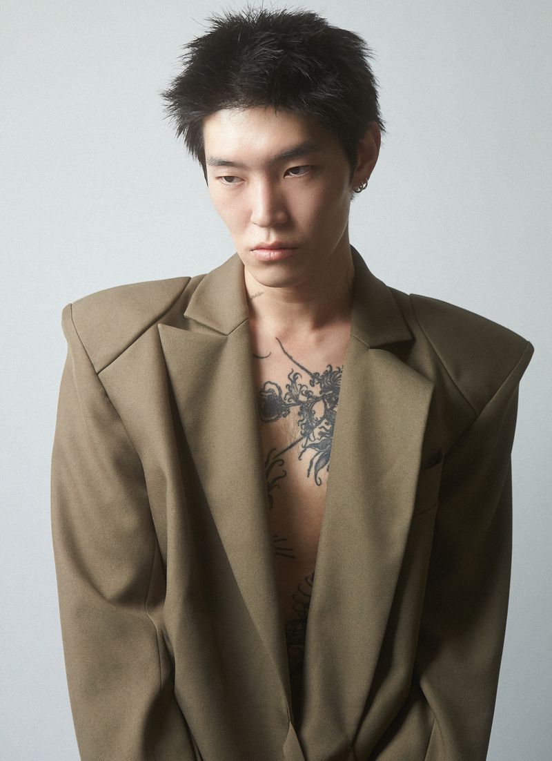 Portrait of an adult male with dark hair and a tattoo on his chest wearing a tan blazer against a neutral background in a photo studio.