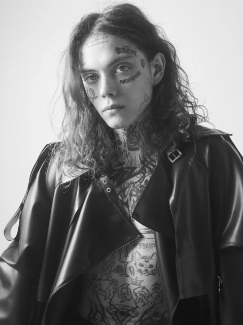 Black and white portrait of a young adult with tattoos posing indoors, wearing a dark coat, showcasing fashion and alternative style.