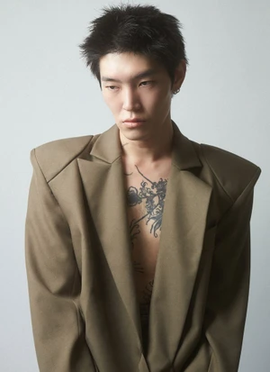 East Asian male model Karl Heinz Hirschberger wearing an oversized structured brown blazer with chest tattoos exposed against a grey backdrop.