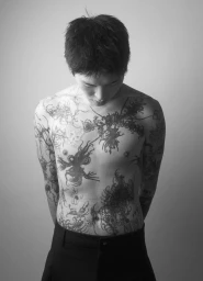 Shirtless model Karl Heinz Hirschberger looking down, showing intricate black tattoos on his chest and stomach against a grey backdrop.