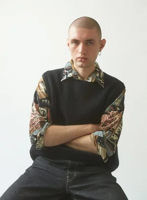 Close-up portrait of a man with a shaved head wearing a black vest and printed shirt against a plain white background