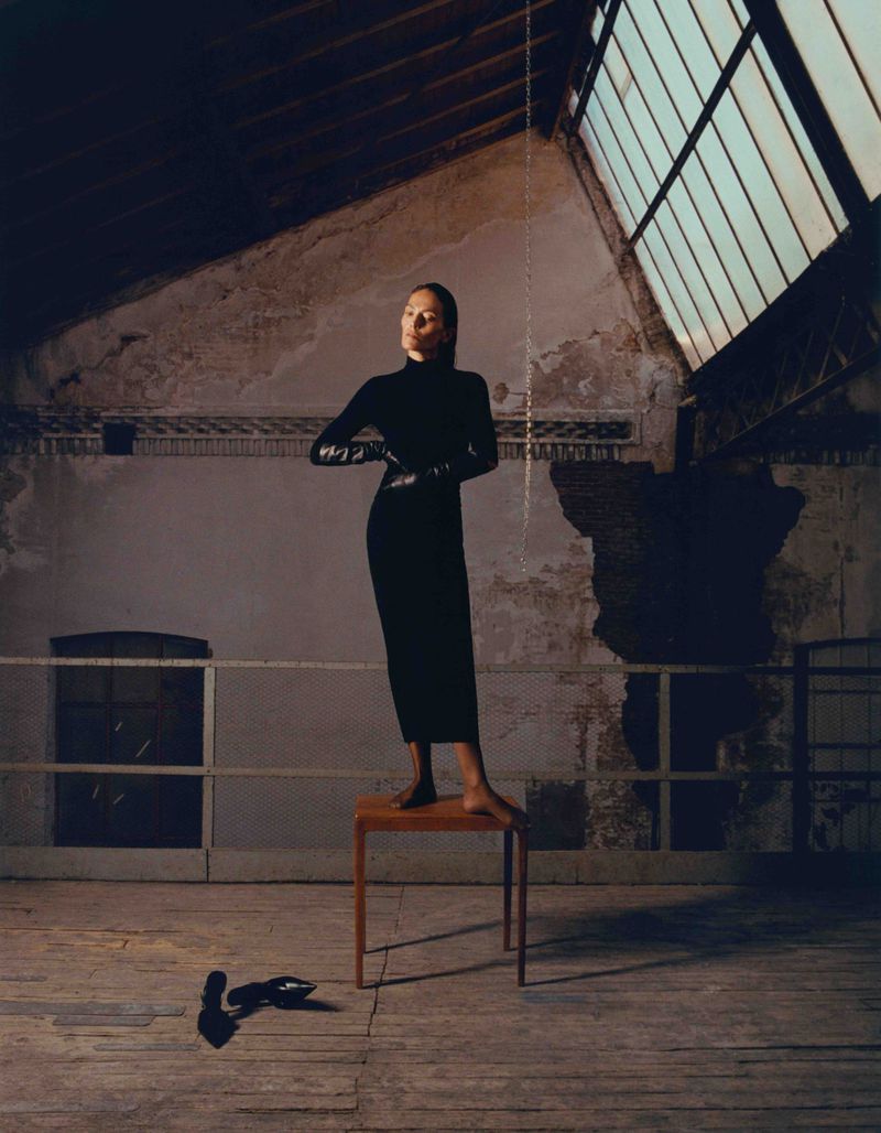 Woman in black dress poses on stool in industrial space with aged walls and natural light, wearing gloves and standing near her shoes.