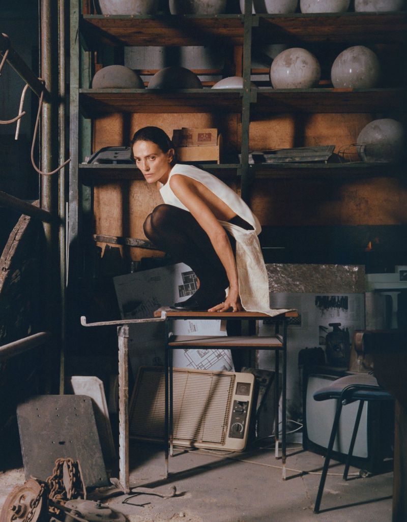 A woman in a white dress crouches on a table in a cluttered indoor space filled with shelves, containers, and various objects.