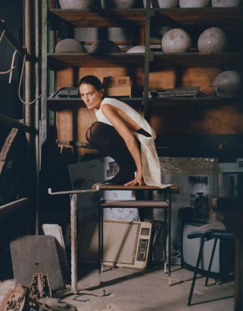 A woman in a white dress crouches on a table in a cluttered indoor space filled with shelves, containers, and various objects.