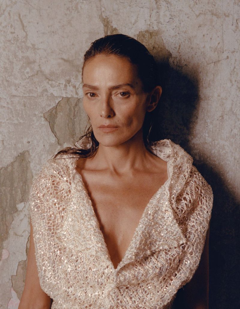 A woman with dark hair and light skin poses in front of a mottled wall, wearing a cream-colored, lace-like garment. She looks straight ahead.