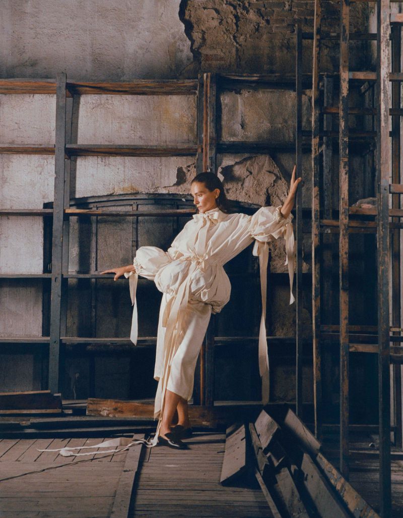 A woman in a billowing cream dress poses artistically in a dilapidated room with exposed brick and metal shelves, lit by natural light.