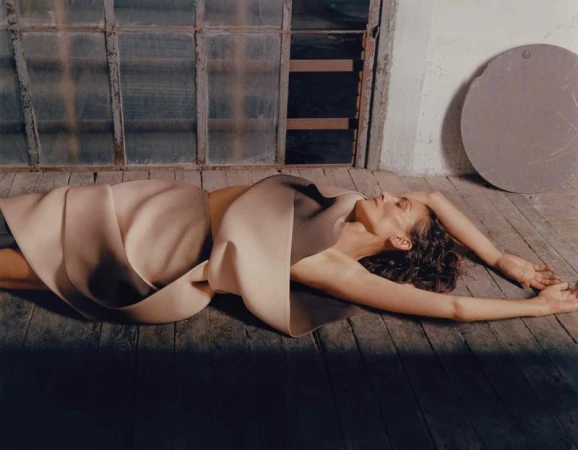 Adult woman in a draped garment lies on wooden planks indoors in front of an old window, illuminated by dim, natural light.