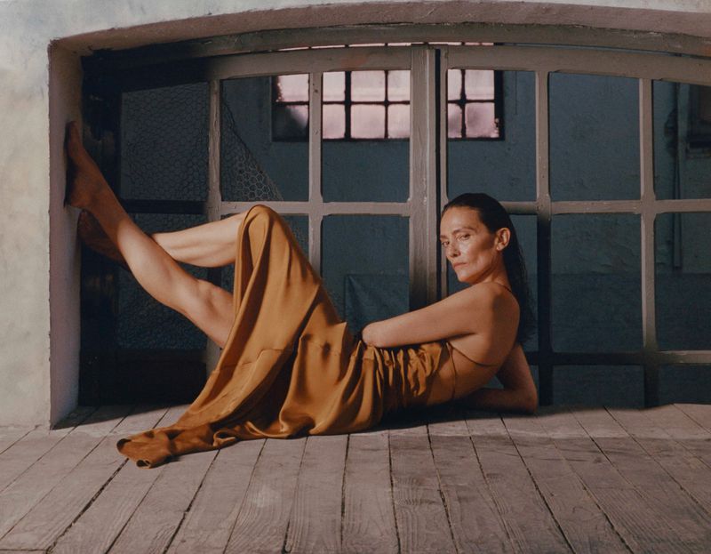 Adult woman in a gold dress leans against a white wall next to a large window with a view of the dusky sunset.