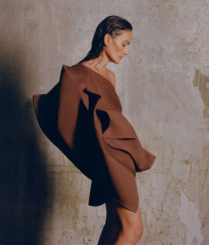 Woman posing in brown sculptural dress against a textured wall. Studio lighting highlights the dress's unique design. Fashion photography.