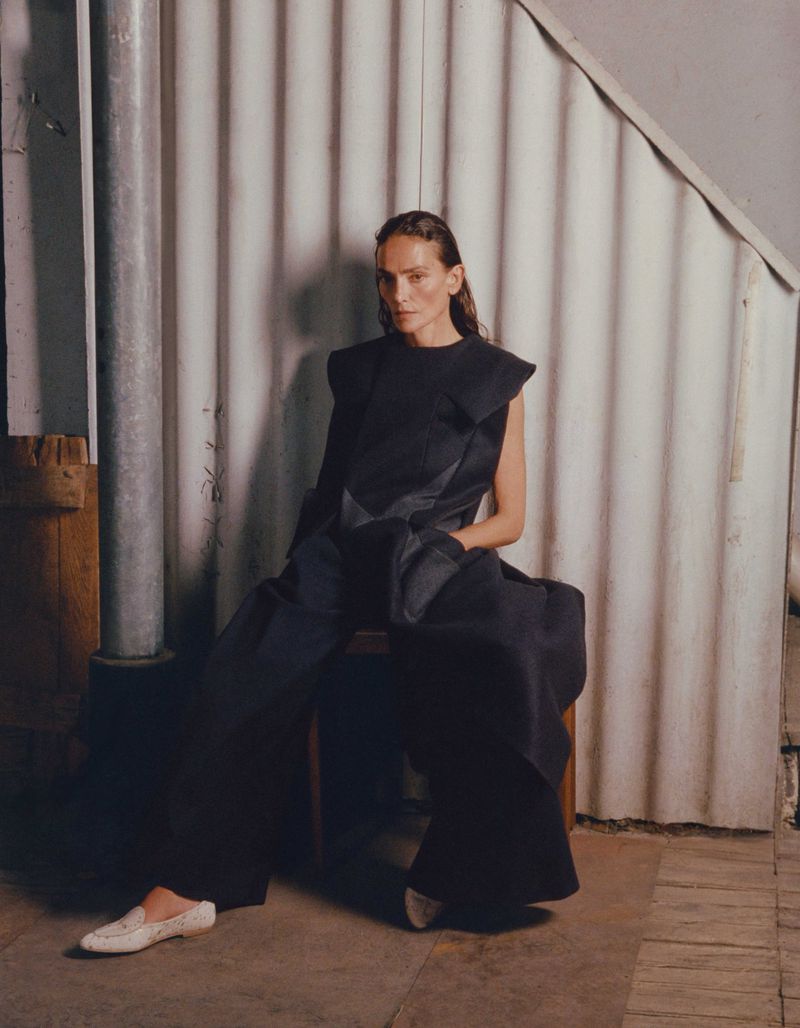 Adult female in a sleeveless black outfit seated on a wooden chair in front of a corrugated metal wall and a metal pipe.